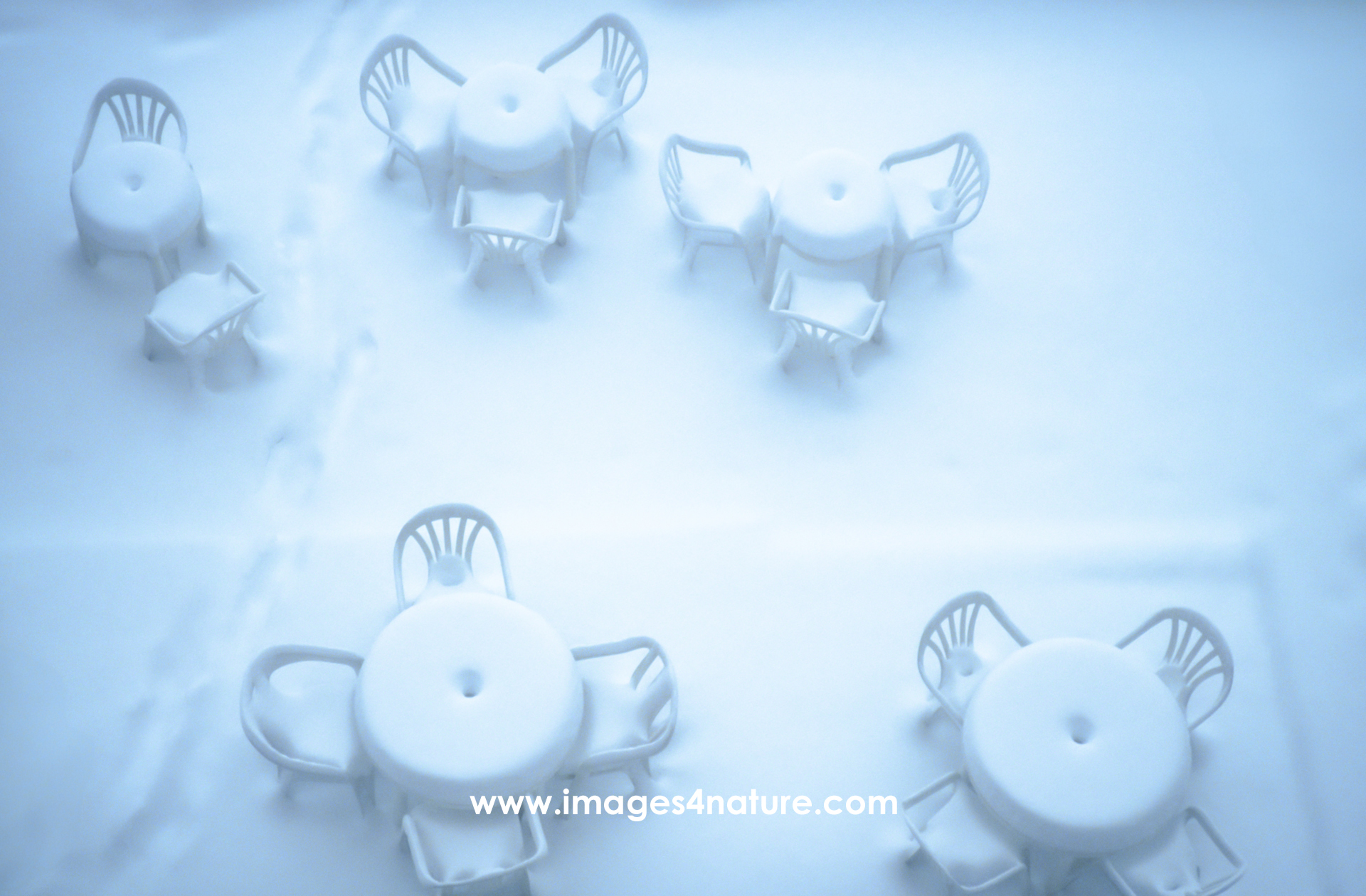 Four white plastic tables and chairs fully covered by fresh snow