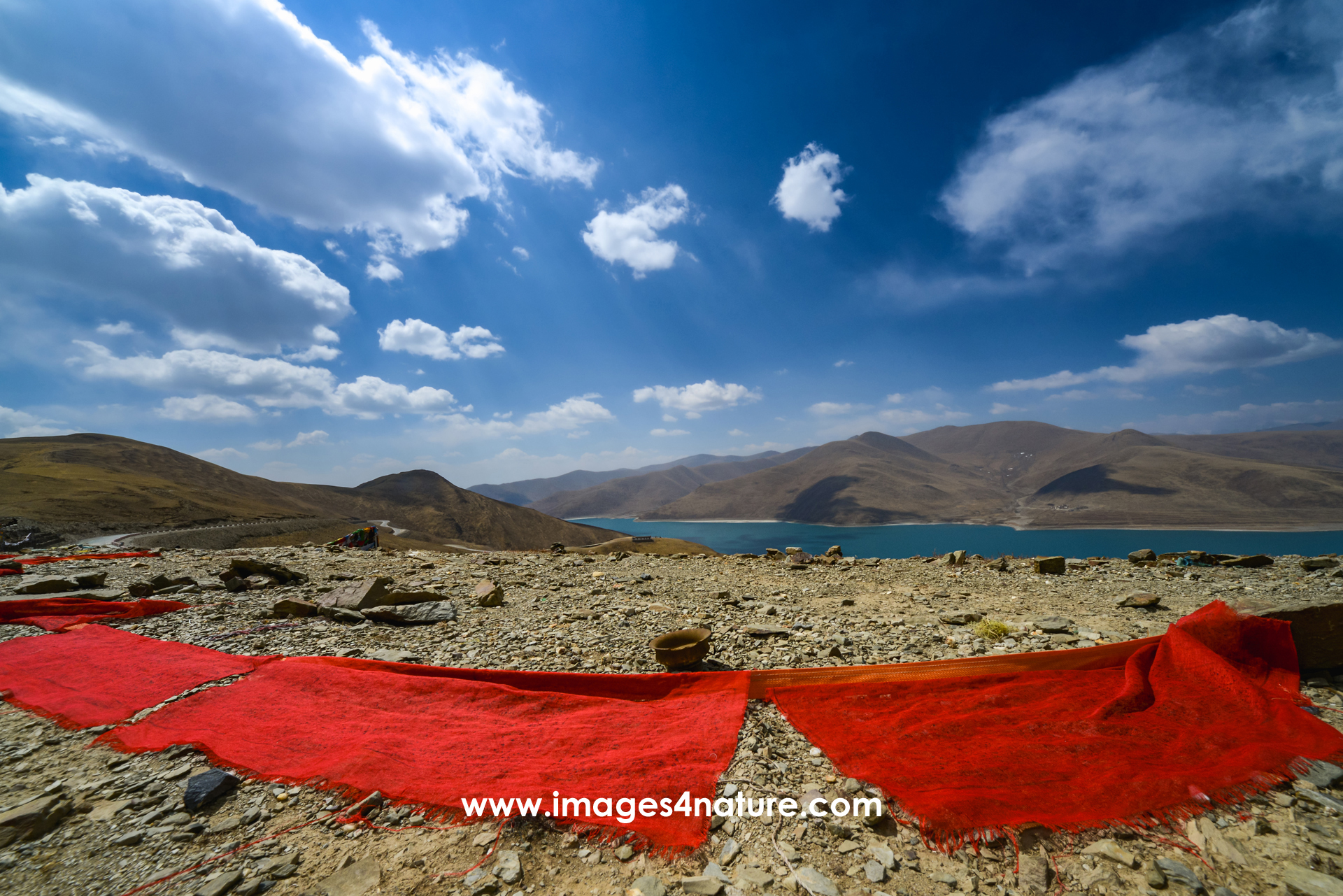 Large dark red prayer flags laid out on the arid floor with mountains and a beautiful lake in the background