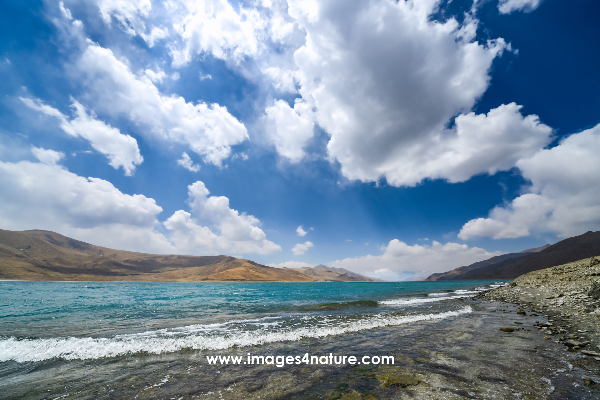 Rocky beach with waves of the Yamzho Yumco lake against blue sky with large white clouds