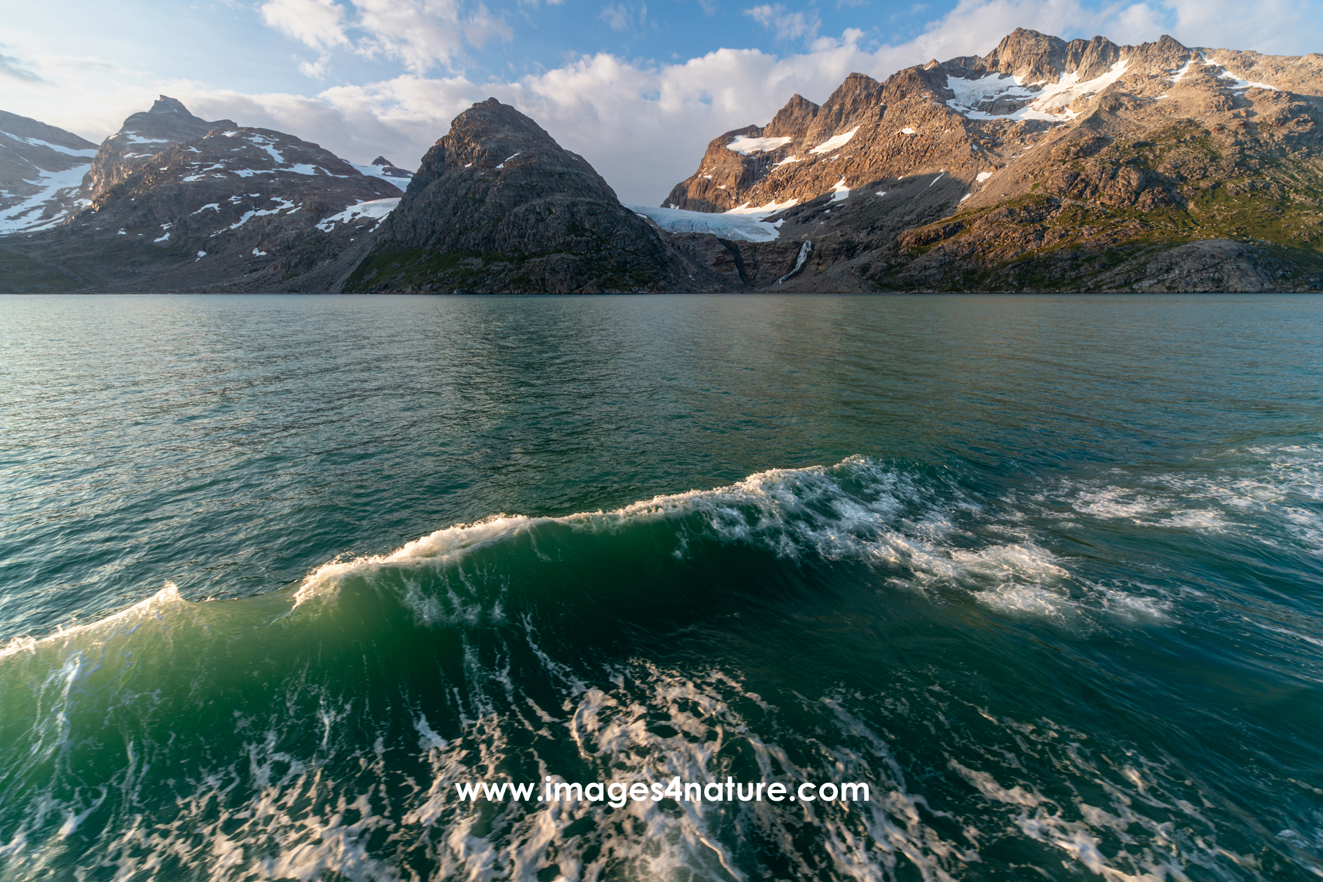 Sunlit bow wave of a cruise ship against the backdrop of snow covered mountains